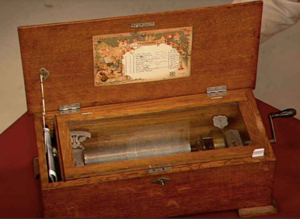 A wooden music box rests with its lid open. Inside are metal hinges and a metal cylinder. Outside the box is a metal handle attached to the metal cylinder which causes the cylinder to rotate. Affixed to the inside of the open lid there is a rectangular plate with a list of music.